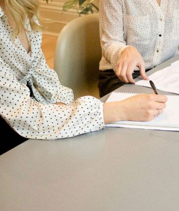 Two people in polka dot blouses sign documents at a table during a meeting.